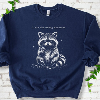 Navy sweatshirt flat lay with a white stippled illustration of a fluffy raccoon sitting up and holding a small mushroom, with small sparkles, below the lowercase typewriter-style words "i ate the wrong mushroom", on white brick with ivy sprigs.