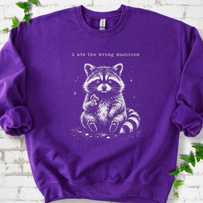 Purple sweatshirt flat lay with a white stippled illustration of a fluffy raccoon sitting up and holding a small mushroom, with small sparkles, below the lowercase typewriter-style words "i ate the wrong mushroom", on white brick with ivy sprigs.