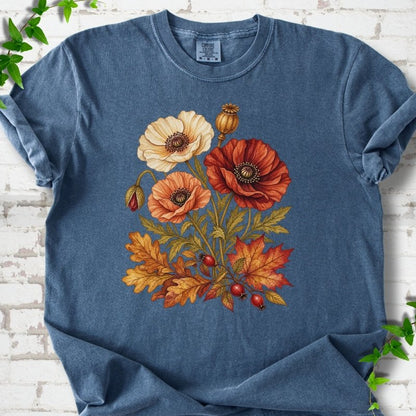 Denim blue short sleeve t-shirt with a botanical bouquet of cream, peach and deep red poppies with a seed pod, oak and maple leaves and red rose hips, printed on the chest, flat lay on white brick with ivy
