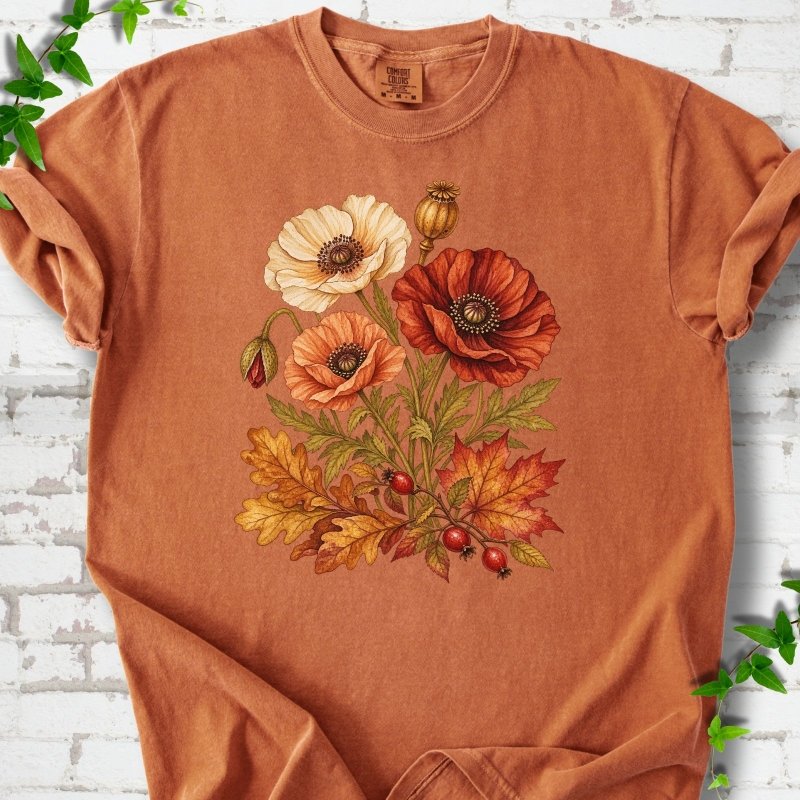 Yam burnt orange short sleeve t-shirt with a botanical bouquet of cream, peach and deep red poppies with a seed pod, oak and maple leaves and red rose hips, printed on the chest, flat lay on white brick with ivy