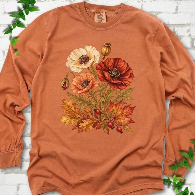 Yam burnt orange long sleeve t-shirt with a botanical bouquet of cream, peach and deep red poppies with a seed pod, oak and maple leaves and red rose hips, printed on the chest, flat lay on white brick with ivy