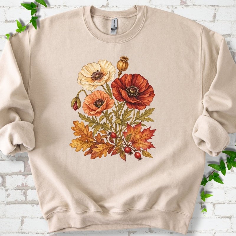 Sand beige crewneck sweatshirt with a botanical bouquet of cream, peach and deep red poppies with a seed pod, oak and maple leaves and red rose hips, printed on the chest, flat lay on white brick with ivy