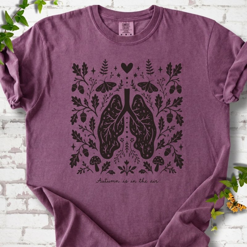 Berry Comfort Colors® T-shirt front mockup featuring a dark, symmetrical folk-botanical graphic centered on stylized human lungs and branching airways. The lungs are framed by leafy branches, acorns, two mushrooms.