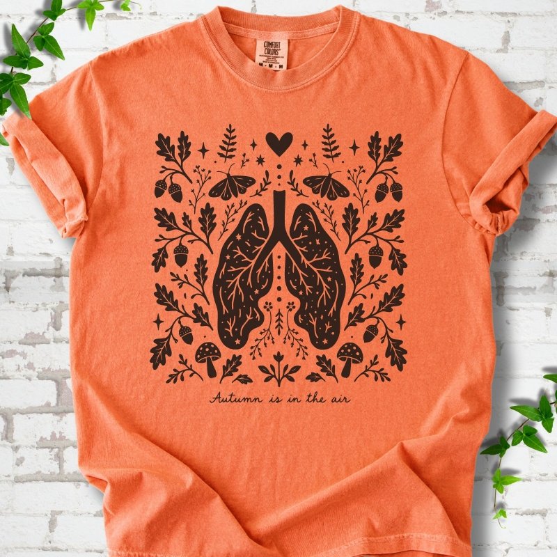Burnt Orange Comfort Colors® T-shirt front mockup featuring a dark, symmetrical folk-botanical graphic centered on stylized human lungs and branching airways. The lungs are framed by leafy branches, acorns, two mushrooms.