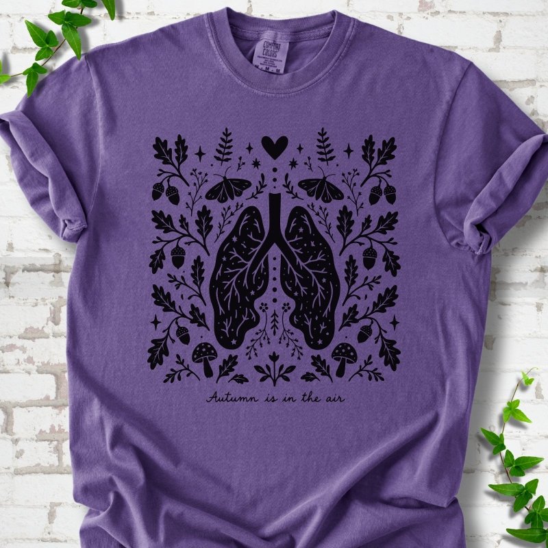 Grape Comfort Colors® T-shirt front mockup featuring a dark, symmetrical folk-botanical graphic centered on stylized human lungs and branching airways. The lungs are framed by leafy branches, acorns, two mushrooms.