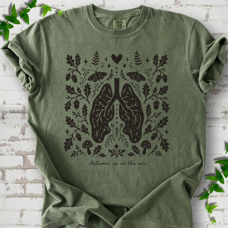 Moss Comfort Colors® T-shirt front mockup featuring a dark, symmetrical folk-botanical graphic centered on stylized human lungs and branching airways. The lungs are framed by leafy branches, acorns, two mushrooms.
