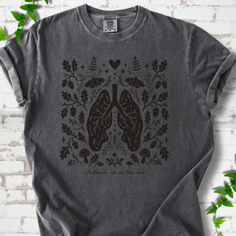 Pepper Comfort Colors® T-shirt front mockup featuring a dark, symmetrical folk-botanical graphic centered on stylized human lungs and branching airways. The lungs are framed by leafy branches, acorns, two mushrooms.