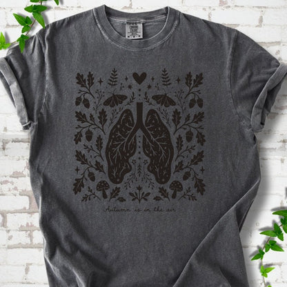 Pepper Comfort Colors® T-shirt front mockup featuring a dark, symmetrical folk-botanical graphic centered on stylized human lungs and branching airways. The lungs are framed by leafy branches, acorns, two mushrooms.