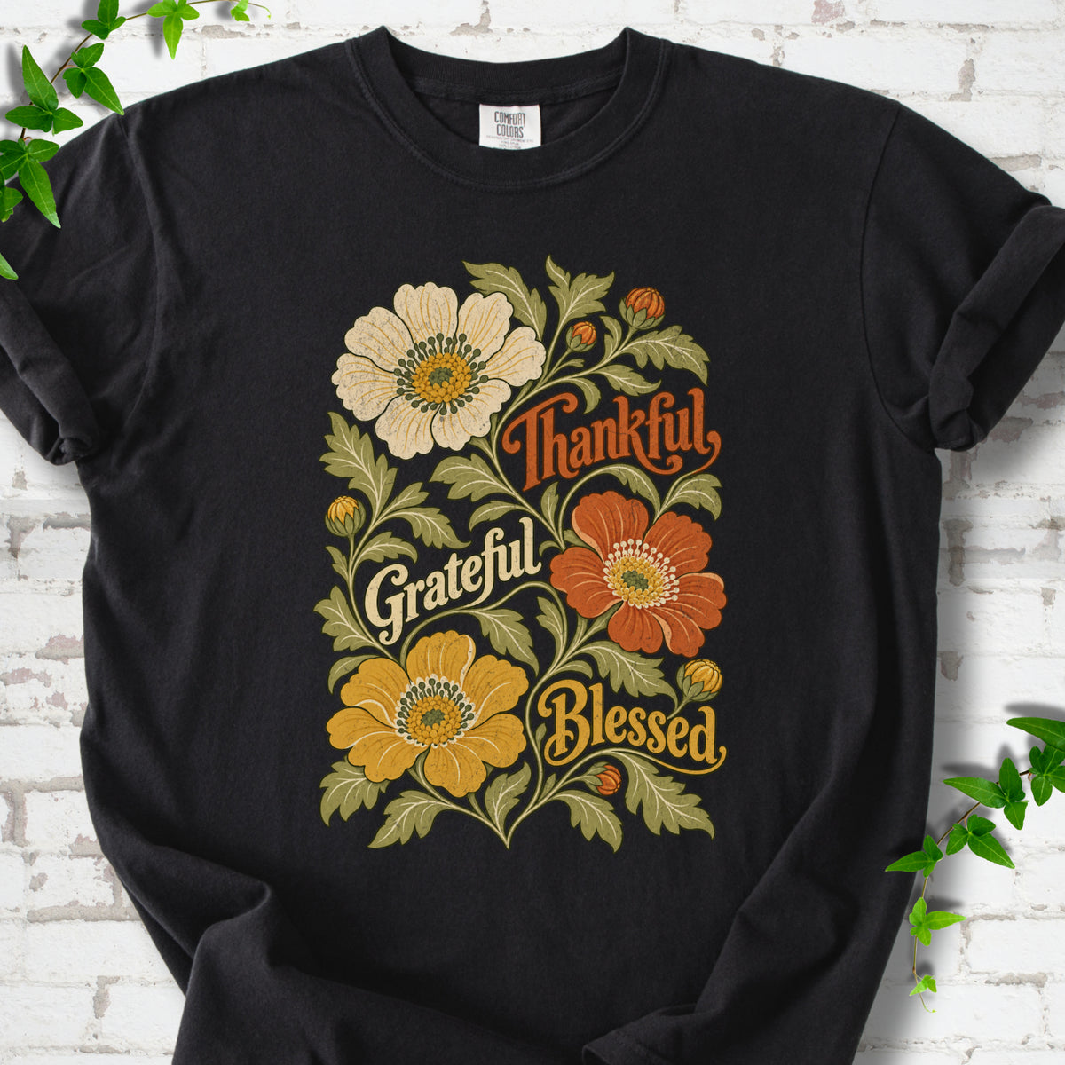 Black T-shirt flat-lay mockup with orange, cream and yellow flowers framing "Thankful, Grateful, Blessed" lettering.