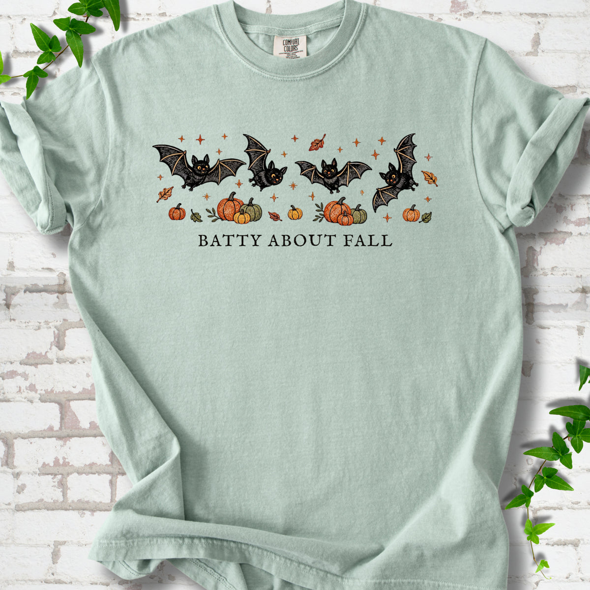 Bay Comfort Colors® T-shirt flat lay featuring four black bats above pumpkins and autumn leaves with “Batty About Fall” lettering.