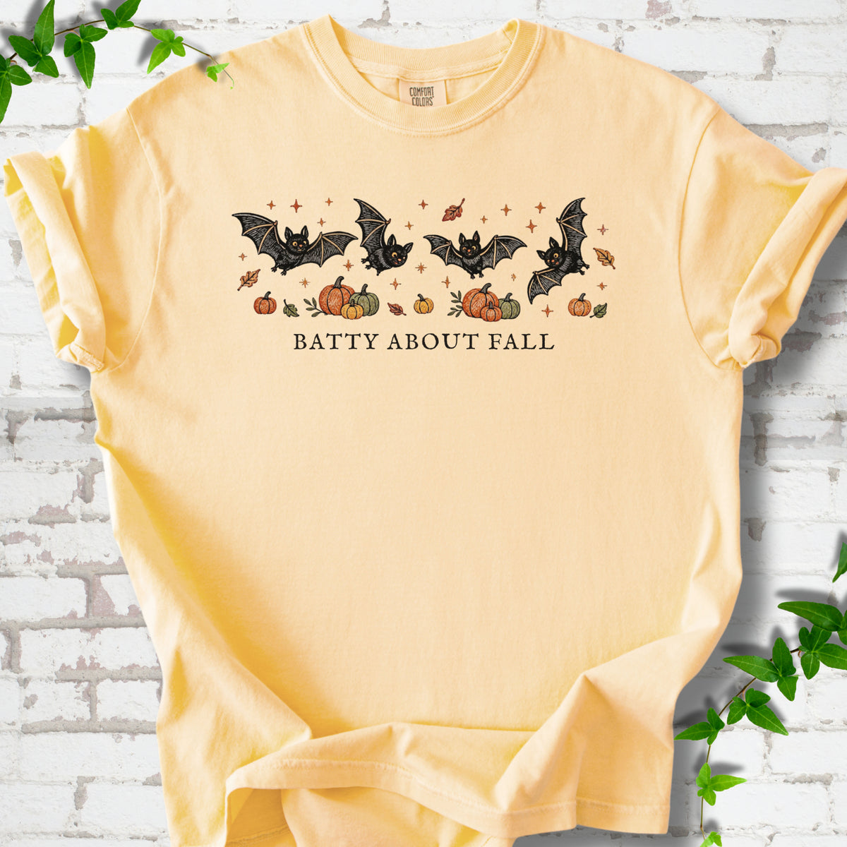 Butter Comfort Colors® T-shirt flat lay featuring four black bats above pumpkins and autumn leaves with “Batty About Fall” lettering.
