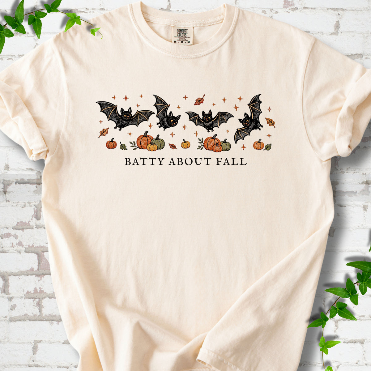 Ivory Comfort Colors® T-shirt flat lay featuring four black bats above pumpkins and autumn leaves with “Batty About Fall” lettering.