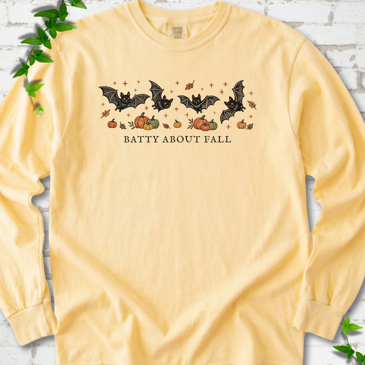 Butter Comfort Colors® long sleeve tee flat lay featuring four black bats above pumpkins and autumn leaves with “Batty About Fall” lettering.