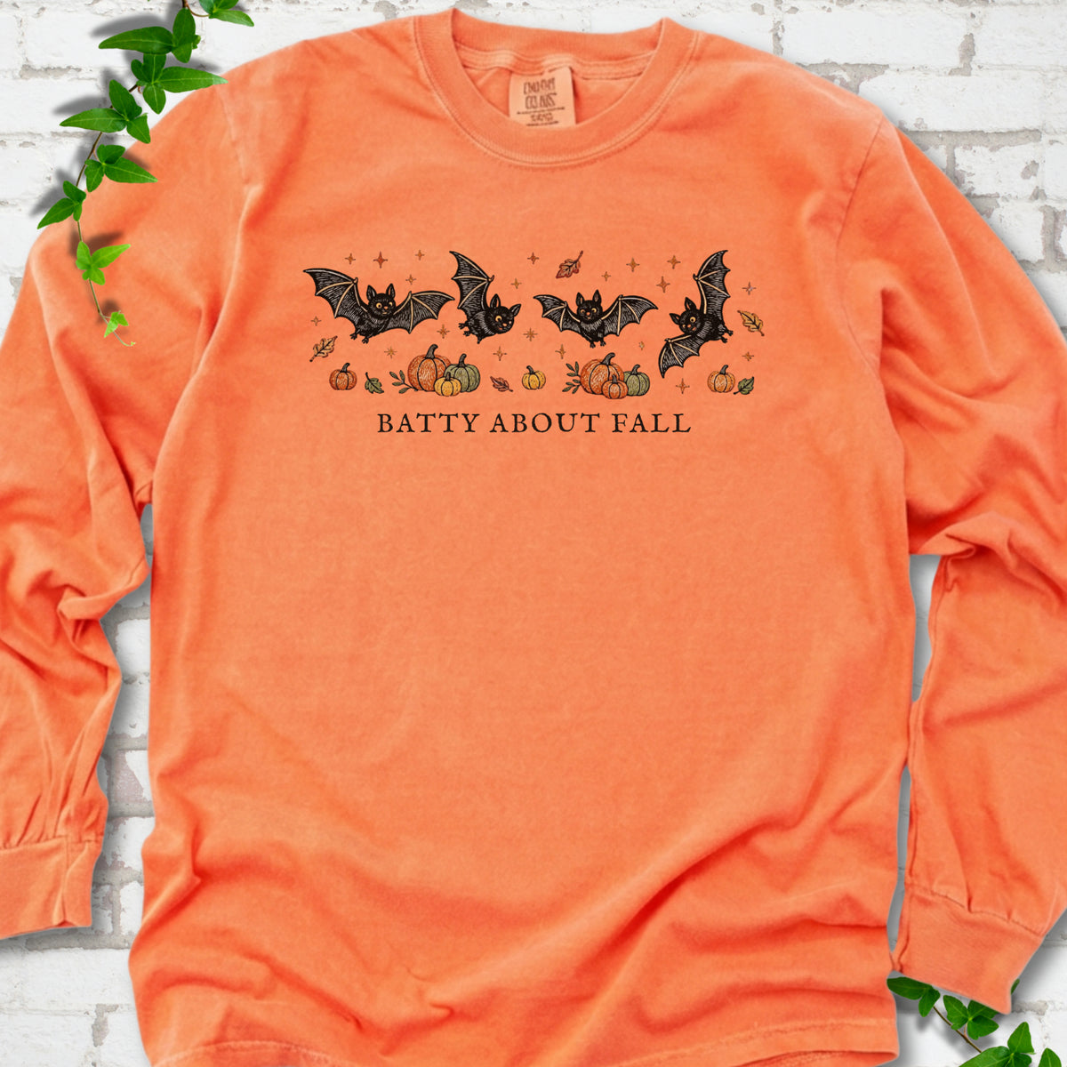 Melon Comfort Colors® long sleeve tee flat lay featuring four black bats above pumpkins and autumn leaves with “Batty About Fall” lettering.