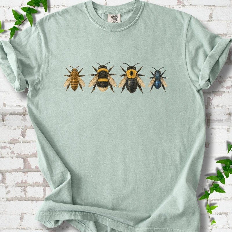 Bay pale sage green short sleeve t-shirt with a row of four bees across the chest: a slim golden honey bee, a fuzzy black and yellow bumble bee, a glossy black bee with a yellow collar and a small metallic blue bee, flat lay on white brick with ivy