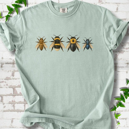 Bay pale sage green short sleeve t-shirt with a row of four bees across the chest: a slim golden honey bee, a fuzzy black and yellow bumble bee, a glossy black bee with a yellow collar and a small metallic blue bee, flat lay on white brick with ivy
