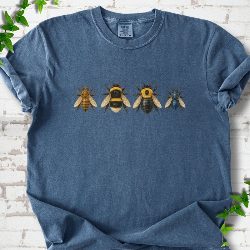 Denim blue short sleeve t-shirt with a row of four bees across the chest: a slim golden honey bee, a fuzzy black and yellow bumble bee, a glossy black bee with a yellow collar and a small metallic blue bee, flat lay on white brick with ivy