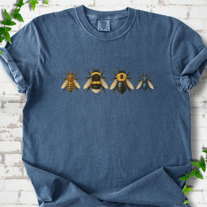 Denim blue short sleeve t-shirt with a row of four bees across the chest: a slim golden honey bee, a fuzzy black and yellow bumble bee, a glossy black bee with a yellow collar and a small metallic blue bee, flat lay on white brick with ivy