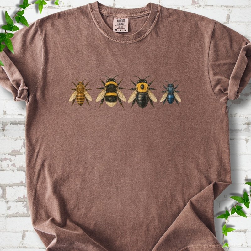Espresso brown short sleeve t-shirt with a row of four bees across the chest: a slim golden honey bee, a fuzzy black and yellow bumble bee, a glossy black bee with a yellow collar and a small metallic blue bee, flat lay on white brick with ivy