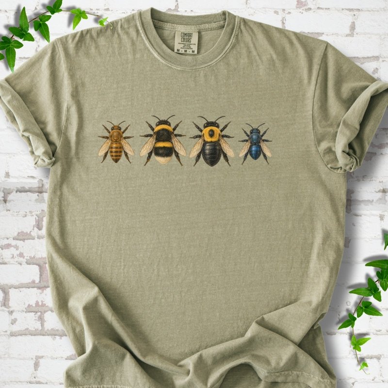 Khaki sage short sleeve t-shirt with a row of four bees across the chest: a slim golden honey bee, a fuzzy black and yellow bumble bee, a glossy black bee with a yellow collar and a small metallic blue bee, flat lay on white brick with ivy