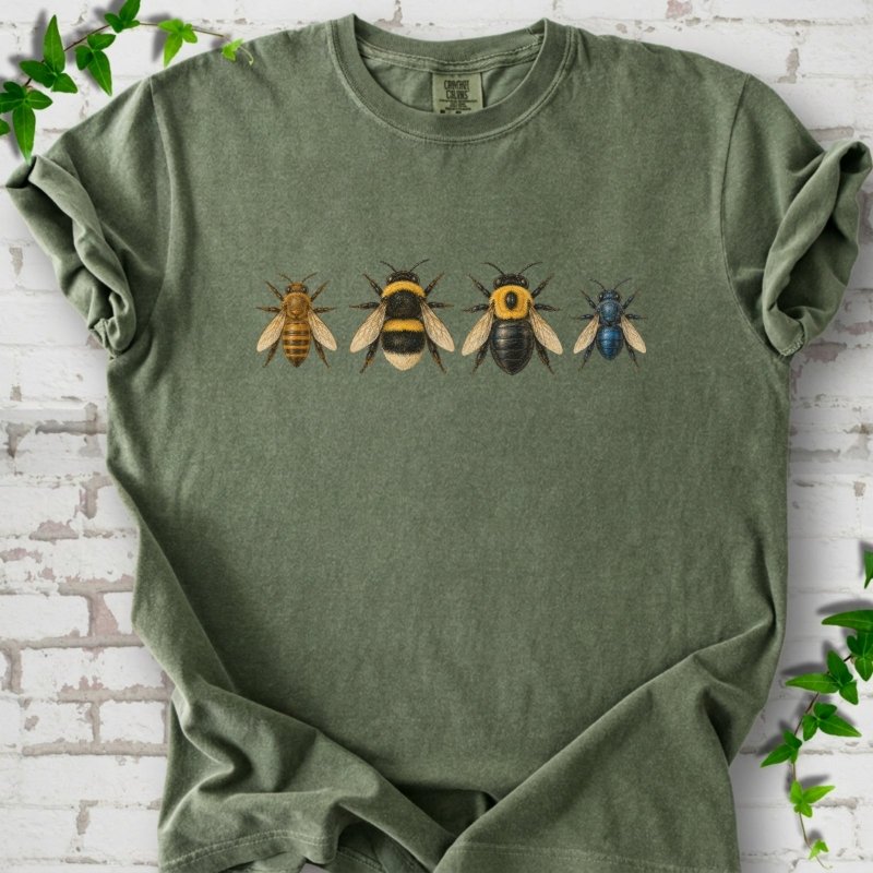 Moss green short sleeve t-shirt with a row of four bees across the chest: a slim golden honey bee, a fuzzy black and yellow bumble bee, a glossy black bee with a yellow collar and a small metallic blue bee, flat lay on white brick with ivy