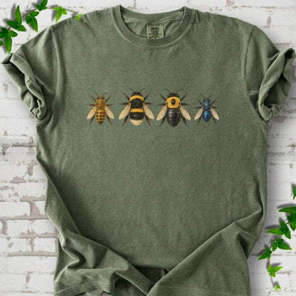 Moss green short sleeve t-shirt with a row of four bees across the chest: a slim golden honey bee, a fuzzy black and yellow bumble bee, a glossy black bee with a yellow collar and a small metallic blue bee, flat lay on white brick with ivy