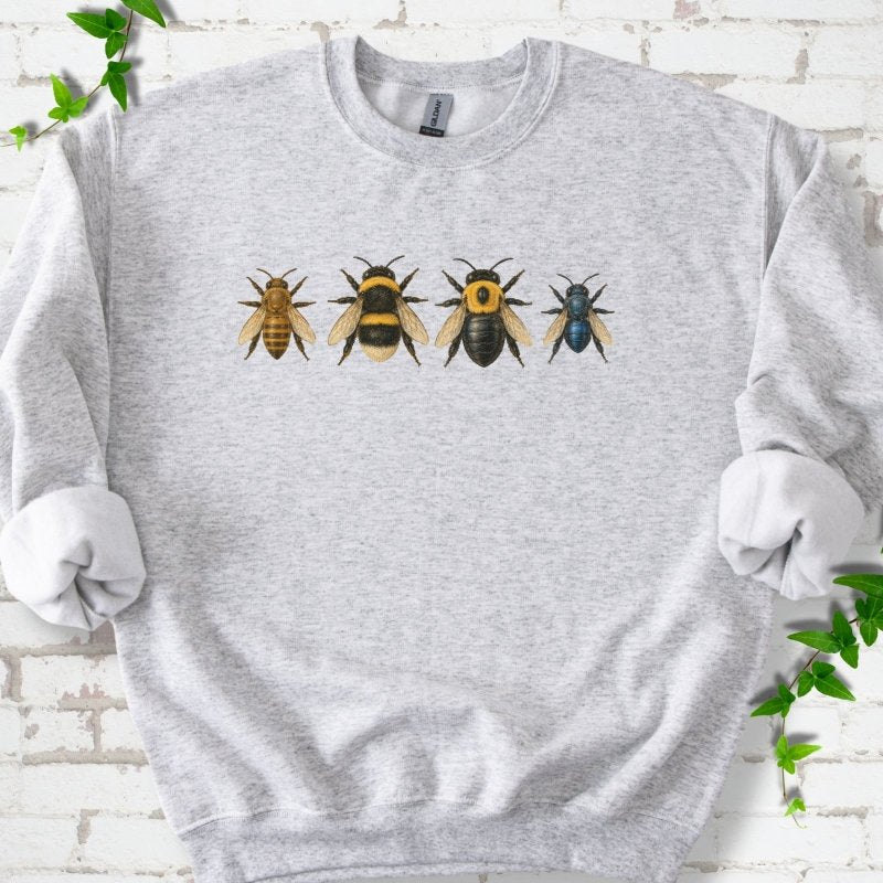 Ash light gray crewneck sweatshirt with a row of four bees across the chest: a slim golden honey bee, a fuzzy black and yellow bumble bee, a glossy black bee with a yellow collar and a small metallic blue bee, flat lay on white brick with ivy