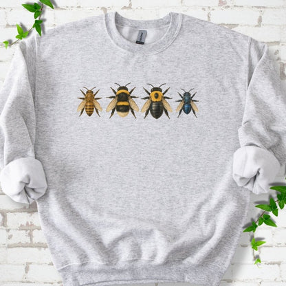 Ash light gray crewneck sweatshirt with a row of four bees across the chest: a slim golden honey bee, a fuzzy black and yellow bumble bee, a glossy black bee with a yellow collar and a small metallic blue bee, flat lay on white brick with ivy