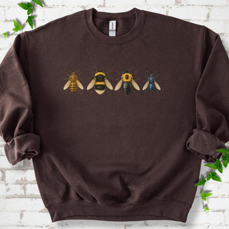 Dark Chocolate brown crewneck sweatshirt with a row of four bees across the chest: a slim golden honey bee, a fuzzy black and yellow bumble bee, a glossy black bee with a yellow collar and a small metallic blue bee, flat lay on white brick with ivy