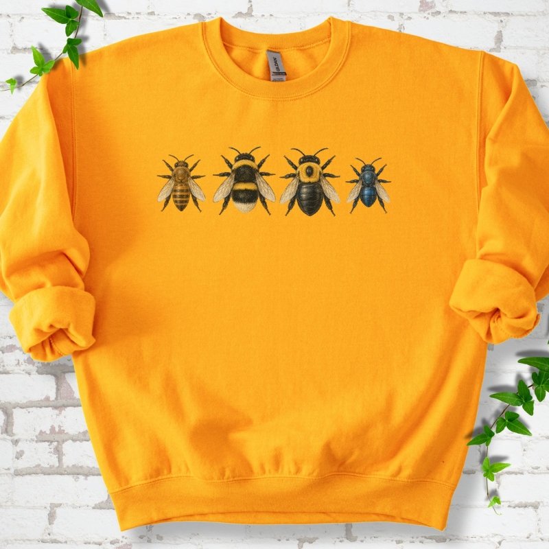 Gold yellow crewneck sweatshirt with a row of four bees across the chest: a slim golden honey bee, a fuzzy black and yellow bumble bee, a glossy black bee with a yellow collar and a small metallic blue bee, flat lay on white brick with ivy
