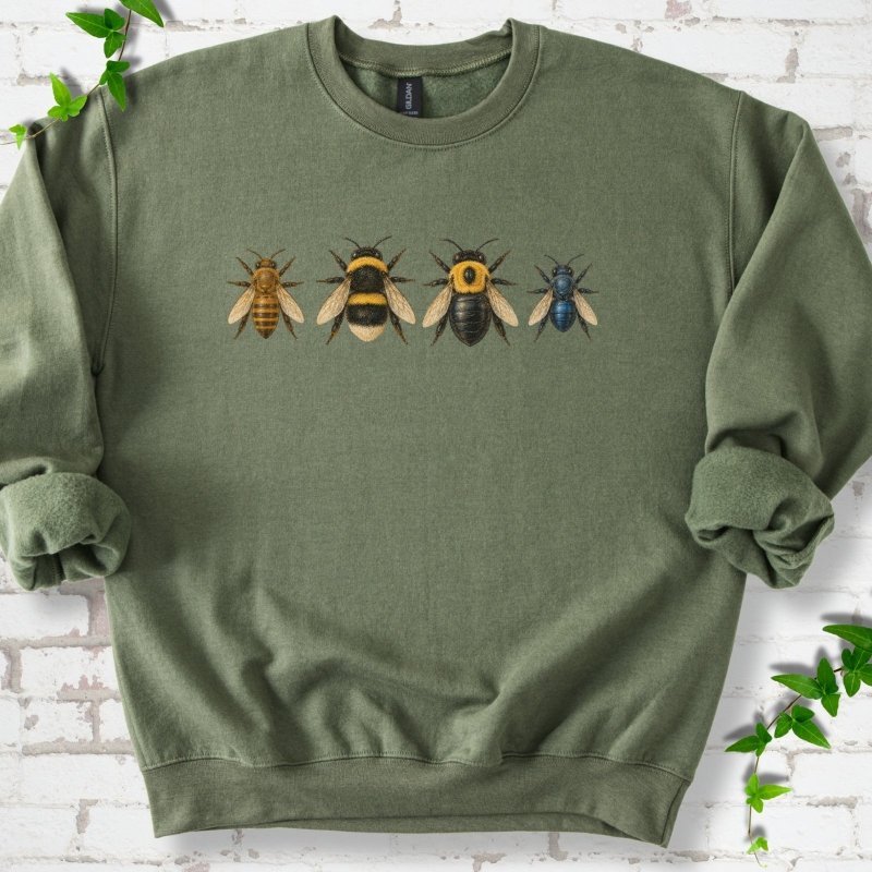 Military olive green crewneck sweatshirt with a row of four bees across the chest: a slim golden honey bee, a fuzzy black and yellow bumble bee, a glossy black bee with a yellow collar and a small metallic blue bee, flat lay on white brick with ivy