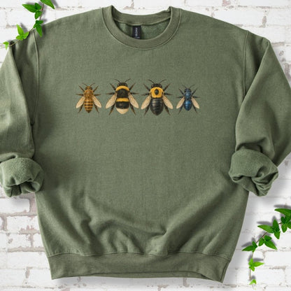 Military olive green crewneck sweatshirt with a row of four bees across the chest: a slim golden honey bee, a fuzzy black and yellow bumble bee, a glossy black bee with a yellow collar and a small metallic blue bee, flat lay on white brick with ivy