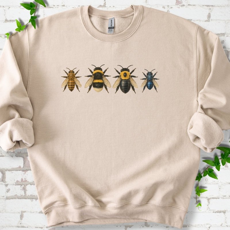 Sand beige crewneck sweatshirt with a row of four bees across the chest: a slim golden honey bee, a fuzzy black and yellow bumble bee, a glossy black bee with a yellow collar and a small metallic blue bee, flat lay on white brick with ivy