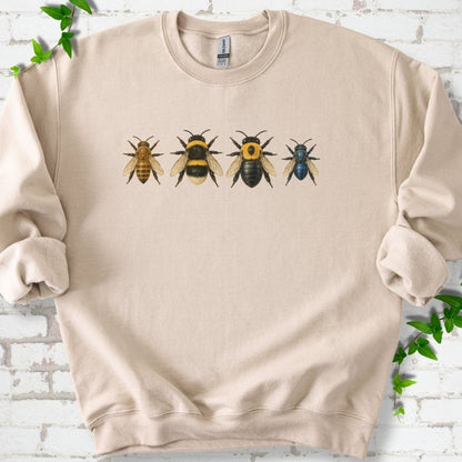 Sand beige crewneck sweatshirt with a row of four bees across the chest: a slim golden honey bee, a fuzzy black and yellow bumble bee, a glossy black bee with a yellow collar and a small metallic blue bee, flat lay on white brick with ivy