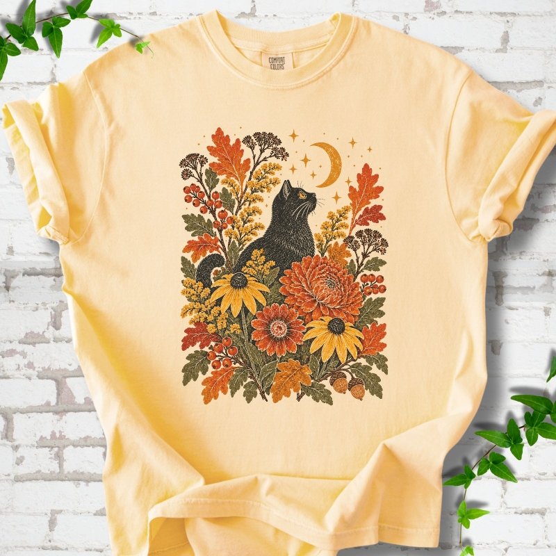 Front flat lay of a Butter short sleeve T-shirt with a black cat beneath a crescent moon among autumn flowers and oak leaves, on white brick with green leafy sprigs.