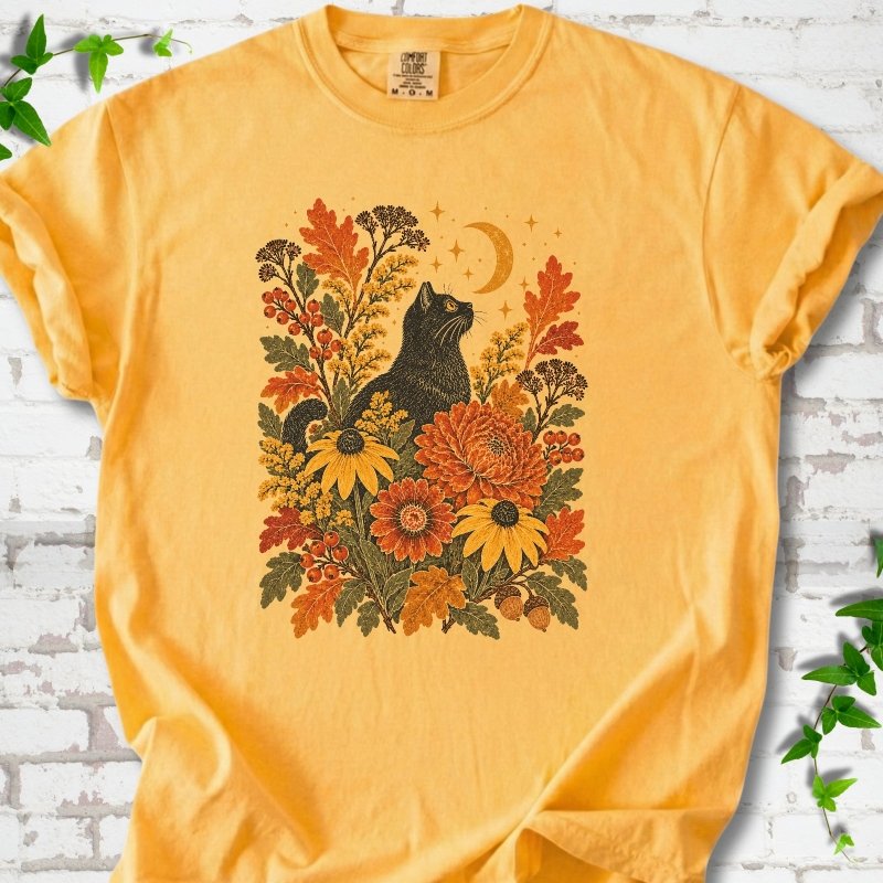 Front flat lay of a Citrus short sleeve T-shirt with a black cat beneath a crescent moon among autumn flowers and oak leaves, on white brick with green leafy sprigs.
