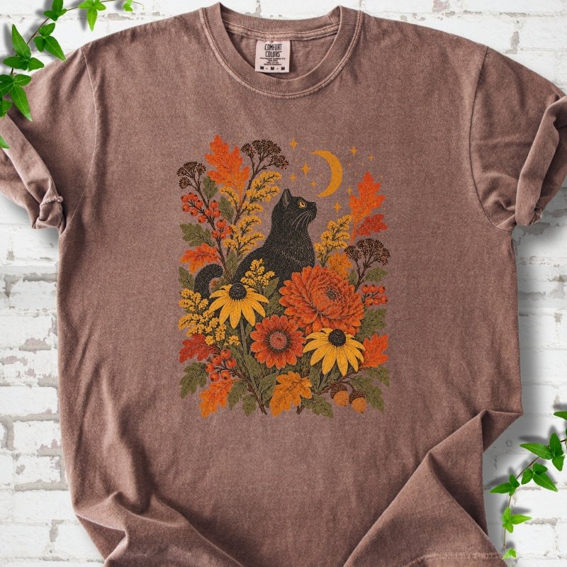 Front flat lay of a Espresso short sleeve T-shirt with a black cat beneath a crescent moon among autumn flowers and oak leaves, on white brick with green leafy sprigs.