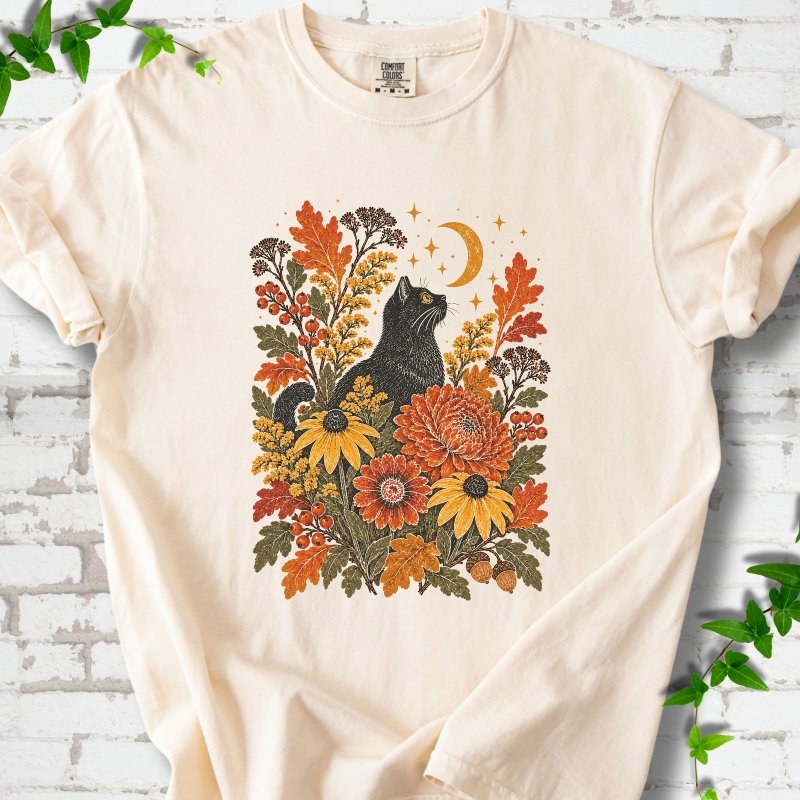 Front flat lay of a Ivory short sleeve T-shirt with a black cat beneath a crescent moon among autumn flowers and oak leaves, on white brick with green leafy sprigs.