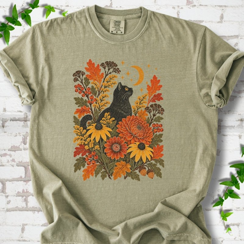 Front flat lay of a Khaki short sleeve T-shirt with a black cat beneath a crescent moon among autumn flowers and oak leaves, on white brick with green leafy sprigs.
