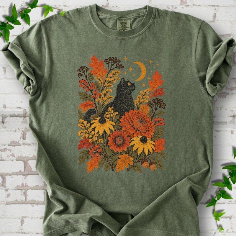 Front flat lay of a Moss short sleeve T-shirt with a black cat beneath a crescent moon among autumn flowers and oak leaves, on white brick with green leafy sprigs.