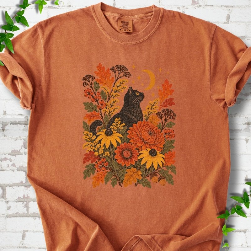Front flat lay of a Yam short sleeve T-shirt with a black cat beneath a crescent moon among autumn flowers and oak leaves, on white brick with green leafy sprigs.