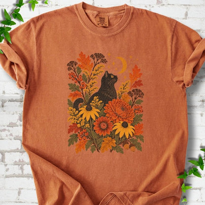 Front flat lay of a Yam short sleeve T-shirt with a black cat beneath a crescent moon among autumn flowers and oak leaves, on white brick with green leafy sprigs.