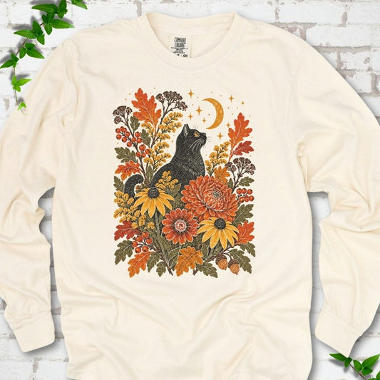 Front flat lay of a Ivory long sleeve T-shirt with a black cat beneath a crescent moon among autumn flowers and oak leaves, on white brick with green leafy sprigs.