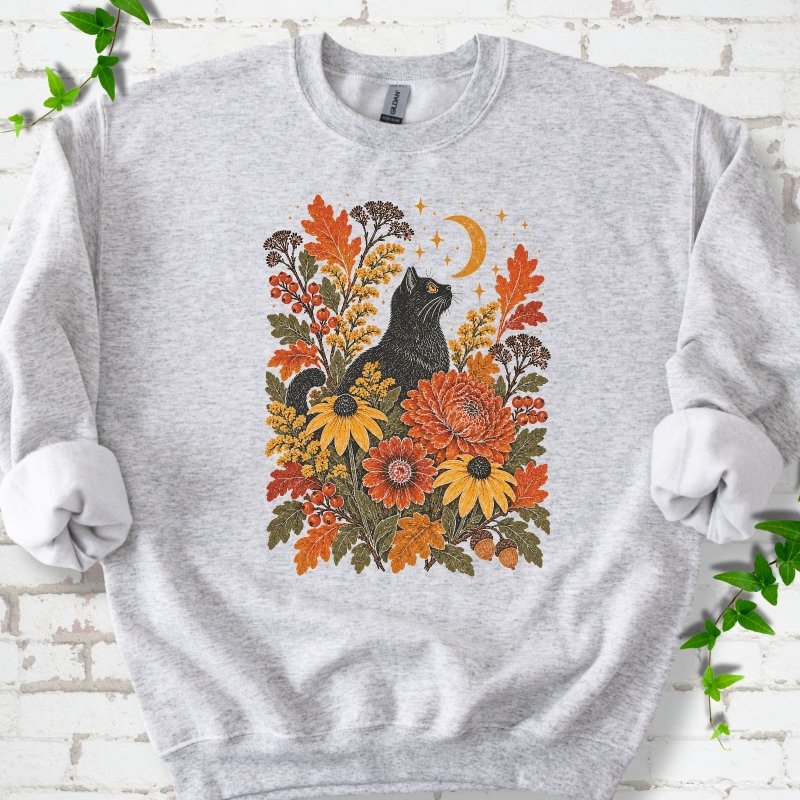 Front flat lay of a Ash crewneck sweatshirt with a black cat beneath a crescent moon among autumn flowers and oak leaves, on white brick with green leafy sprigs.
