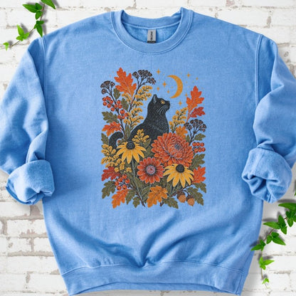 Front flat lay of a Carolina Blue crewneck sweatshirt with a black cat beneath a crescent moon among autumn flowers and oak leaves, on white brick with green leafy sprigs.