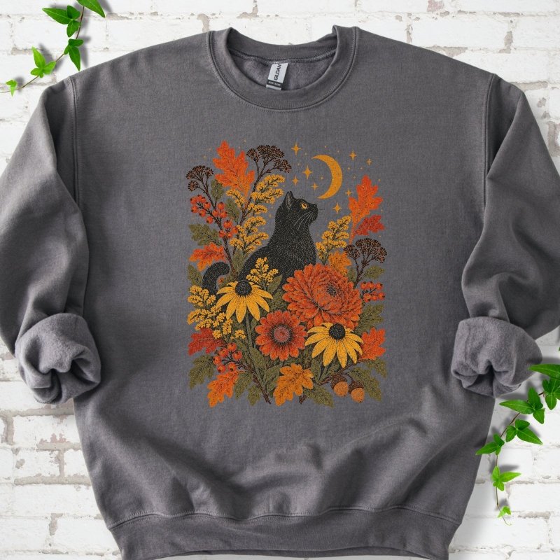 Front flat lay of a Charcoal crewneck sweatshirt with a black cat beneath a crescent moon among autumn flowers and oak leaves, on white brick with green leafy sprigs.