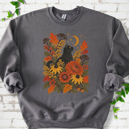 Front flat lay of a Charcoal crewneck sweatshirt with a black cat beneath a crescent moon among autumn flowers and oak leaves, on white brick with green leafy sprigs.