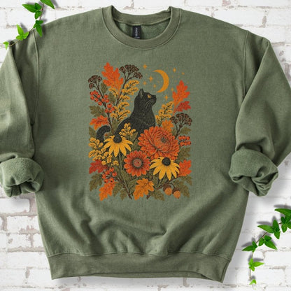 Front flat lay of a Military Green crewneck sweatshirt with a black cat beneath a crescent moon among autumn flowers and oak leaves, on white brick with green leafy sprigs.