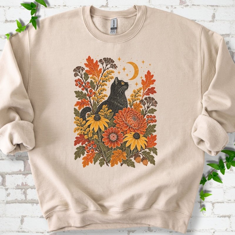 Front flat lay of a Sand crewneck sweatshirt with a black cat beneath a crescent moon among autumn flowers and oak leaves, on white brick with green leafy sprigs.