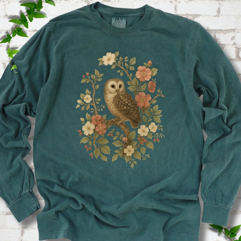 Blue Spruce long sleeve T-shirt flat lay with a botanical illustration of a brown owl perched on a branch, framed by wild roses, cream blossoms, rosehips and olive-green leaves, on white brick with ivy.